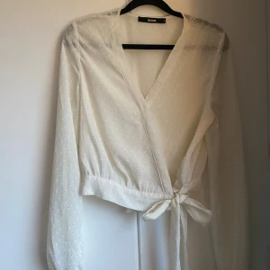 Vit omlottblus med knyt från BikBok - Säljer en vit omlottblus från BikBok med knyt i midjan och diskret prickigt mönster. 