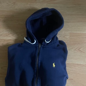 Mörkblå hoodie från Polo Ralph Lauren - Säljer en mörkblå hoodie från Polo Ralph Lauren med dragkedja och huva. Tröjan har det klassiska gula logotrycket på bröstet och är tillverkad i mjuk bomull. Perfekt för en avslappnad stil.