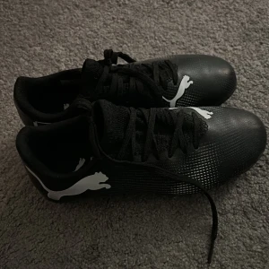 Svarta fotbollsskor från Puma - Säljer ett par svarta fotbollsskor från Puma med vit logga på sidan. Skorna har snörning och mönstrad sula med dobbar för bra grepp på planen. Använda 2 ggr 