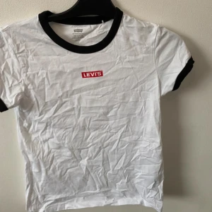 Vit t-shirt med svart krage från Levi's - Vit t-shirt från Levi's med svart krage och svarta ärmslut. Klassisk röd Levi's-logga på bröstet. T-shirten har en enkel och stilren design, perfekt till jeans eller shorts. Frakt tillkommer