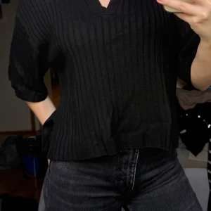 Svart ribbad polotopp - Säljer en svart ribbad polotopp från Stockh LM. Tröjan har korta ärmar, v-ringning och krage. Modellen är croppad och har en avslappnad passform. Perfekt till jeans eller kjol för en stilren look.