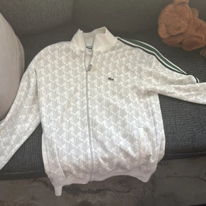 Vit track jacket med mönster och svarta ränder - Säljer en vit track jacket från Lacoste med diskret geometriskt mönster och klassiska svarta ränder på axlarna. Jackan har dragkedja framtill, ribbade muddar och lång ärm. Perfekt för en sportig och stilren look.