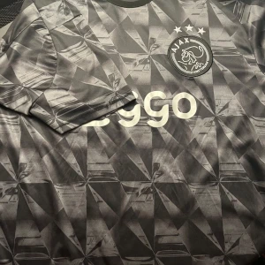 Ajax svart/grå fotbollströja från Adidas - Säljer en svart och grå Ajax fotbollströja från Adidas med geometriskt mönster, vita detaljer och Ziggo-logga på bröstet. Tröjan har korta ärmar och tre vita ränder på axlarna. Perfekt för fotbollsälskare!