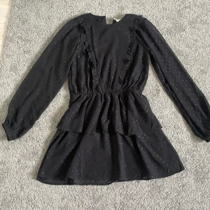 Svart volangklänning med prickigt mönster från Gina Tricot - Supersöt svart klänning från Gina Tricot med små glitter prickar och volangdetaljer framtill. Klänningen har lång ärm, markerad midja och dubbla volanger i kjolen för extra volym. Perfekt för dig nyår, jul och festligt tillfällen 🥰