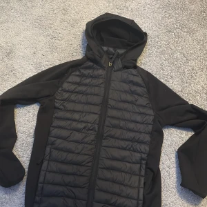 Svart Cardigan från McKinley - Svart jacka från McKinley med quiltad front och rygg samt släta ärmar och sidor. Jackan har huva och hel dragkedja framtill. Perfekt för lager-på-lager och har sportig look.