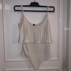 Vit ribbad body från YAS - Säljer en vit ribbad body från YAS i storlek M. Bodyn har smala axelband och ett volangliknande lager över bysten för en trendig look. 