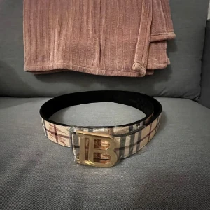 Stiligt burberry bälte.. - Snyggt bälte med rutigt mönster i beige, svart och rött. Bältet har ett stort guldspänne med bokstäver framtill. Perfekt accessoar för att lyfta din outfit.