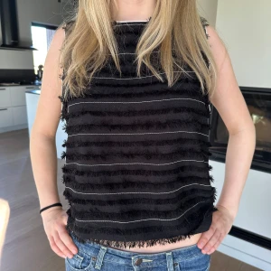 Svart Zara topp - Säljer en svart ärmlös topp med horisontella fransar och tunna vita linjer på framsidan. Toppen har en rak passform och är tillverkad i ett mjukt material. Perfekt för att ge outfiten lite edge! Storlek L men passar mig som vanligtvis har S/M