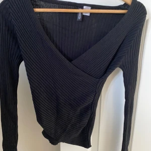 Svart omlottopp från H&M Divided - Säljer en svart ribbad omlottopp från H&M Divided i storlek XS. Toppen har lång ärm och djup v-ringning, perfekt för att styla med jeans eller kjol. Materialet är stretchigt och sitter snyggt på kroppen.