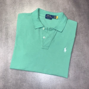 Ralph Lauren piké - Snygg grön pikétröja från Polo Ralph Lauren i klassisk modell. Tröjan har korta ärmar, krage och knäppning med två knappar framtill. Ikoniska broderade loggan i vitt på bröstet. Perfekt för en stilren look. Tröjan är i storlek M alltså passar den någon som är ungefär 165 cm lång. Tröjan är i bra skick och det är bara att höra av er vid funderingar!