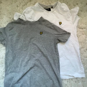 Grå och vit t-shirt från Lyle & Scott - Två klassiska t-shirts från Lyle & Scott, en i grått och en i vitt. Båda har korta ärmar och den ikoniska gula loggan broderad på bröstet. Perfekta basplagg med normal passform och rund halsringning.