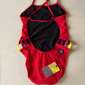 Röd baddräkt från Jolyn - Säljer en röd baddräkt från Jolyn med smala axelband och snygga gula detaljer vid höfterna. Baddräkten har en logga på bröstet och är tillverkad i polyester. Perfekt för simträning eller strandhäng! Endast prövad och har lappen kvar! Skriv gärna om du är intresserad! Eftersom att den är helt ny och limited edition kommat jag sälja den för minst 600 kr.🩷(säljs pga att den var för liten och att den inte gick att lämna tillbaka)