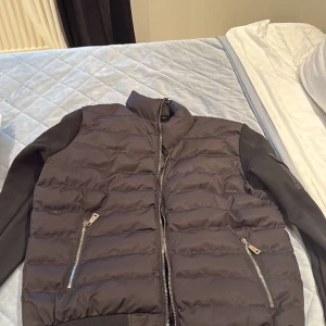 Moncler - Säljer en svart pufferjacka från Moncler med quiltad front och ärmar i stickad bomull. Jackan har två fickor med dragkedja och en Moncler-logga på ärmen. Perfekt för dig som vill ha en stilren och sportig look.