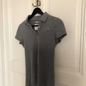 Grå piké t-shirt från Hollister - Grå piké t-shirt från Hollister med klassisk krage och knappslå. Liten broderad logga i vinrött på bröstet. Stretchigt bomullsmaterial och korta ärmar. Perfekt för en avslappnad och stilren look.