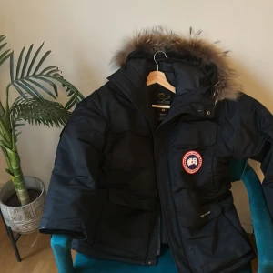 Svart dunjacka med pälskrage från Canada Goose - Säljer en svart dunjacka från Canada Goose med klassisk pälskantad huva och tydlig logotyp på ärmen. Jackan har lång ärm, dragkedja och är perfekt för kalla vinterdagar. Snygg och funktionell design med flera fickor.