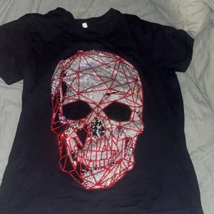 Svart t-shirt med glittrig dödskalle - Cool svart t-shirt med en stor glittrig dödskalle framtill i silver och rött. T-shirten har normal passform och korta ärmar. Perfekt för dig som vill sticka ut med en unik och edgy stil.