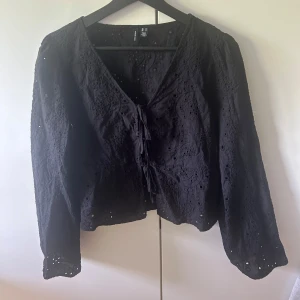 Svart blus med knyt från Vero Moda - Svart blus från Vero Moda med broderade detaljer och knyt framtill. Blusen har lång ärm och v-ringning, samt ett fint hålmönster i tyget som ger en romantisk känsla. Storlek M