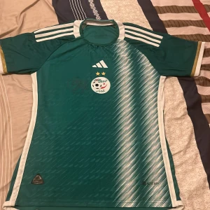 Grön Algeriet fotbollströja från Adidas - Säljer en grön Algeriet fotbollströja från Adidas med vita detaljer och ränder på axlarna. Tröjan har korta ärmar, två stjärnor ovanför landslagets emblem och ett mönster i vitt på framsidan. Perfekt för fotbollsfans!