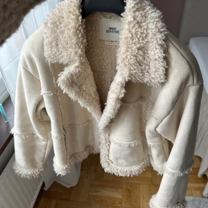 Beige teddyjacka från Urban Outfitters - Säljer en beige teddyjacka från Urban Outfitters med fluffig krage och detaljer längs ärmslut och nederkant. Jackan har en oversized passform och är perfekt för kyligare dagar. Stängs framtill och har en mysig känsla.