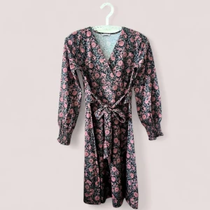 Blommig omlottklänning med volangärm - Supersöt klänning med svart botten och rosa blommor från Three Sixty Five, omlottmodell med v-ringning och knyt i midjan. Långa ärmar med volang och smock vid ärmslut. Perfekt för dig som gillar romantiska detaljer och blommigt.