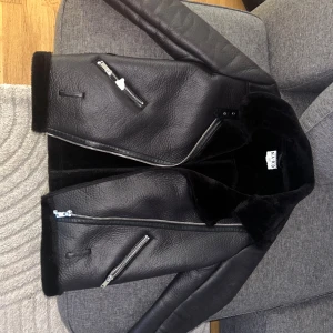 Svart skinnjacka med dragkedjor från NA-KD - Säljer en svart skinnjacka från NA-KD med silvriga dragkedjor och bred krage. Jackan har två snedställda fickor med dragkedja och en mjuk insida. Perfekt för dig som vill ha en cool och stilren look.