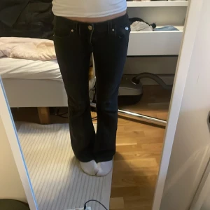 Svarta bootcut jeans - Säljer ett par svarta bootcut jeans med klassisk femficksdesign och silverfärgade knappar. Jeansen har låg midja och är tillverkade i ett stretchigt denimtyg som sitter snyggt över höfterna och låren innan de går ut nertill.
