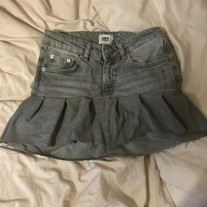 Grå volangkjol i jeans från 157 - Grå jeanskjol med volangkant från 157. Kjolen har klassiska jeansfickor fram och bak, synliga sömmar och rå kant nertill för en edgy look. Perfekt för dig som gillar streetstyle och vill sticka ut med en unik twist på denim.