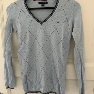 Ljusblå v-ringad tröja Tommy Hilfiger - Ljusblå v-ringad tröja från Tommy Hilfiger med diskret rutig struktur och små logodetaljer. Långa ärmar och ribbade muddar med mörkblå och vita ränder. Perfekt för dig som gillar klassisk och stilren look.