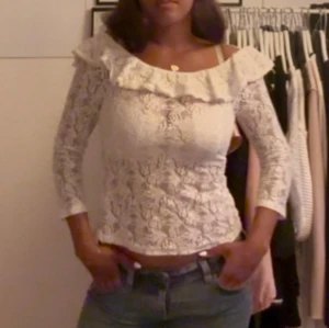 Vit spetsblus med volang och öppen rygg - Superfin vit spetsblus med volangdetalj längs den vida ringningen och snygg öppen rygg. Blusen har trekvartsärm och är figurnära i modellen. Perfekt för dig som gillar romantisk och feminin stil.