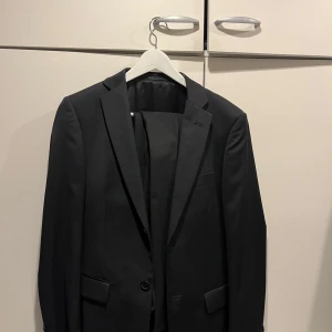 Kavaj och byxor (kostym) - En stilren svart kostym med kavaj och byxor. Kostymen är köpt som ny och har användts en gång. Kavajen har storleken 46 regular slim fit, byxorna 30/32 slim fit.