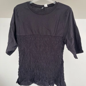 Svart topp med rynkad midja - Svart topp med rund halsringning och halvlånga ärmar. Nedre delen av toppen är rynkad och avslutas med en lätt volangkant. Perfekt för en stilren och trendig look.