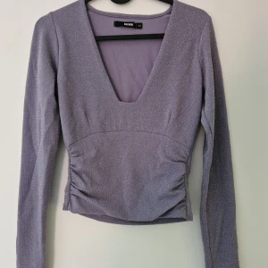 Ljuslila glittrig topp från Bik Bok - Säljer en ljuslila glittrig topp från Bik Bok med djup v-ringning och långa ärmar. Toppen är figurnära och har en croppad passform, perfekt för att lyfta din outfit med lite extra glitter. Finns några tråddragningar likt den på sista bilden.