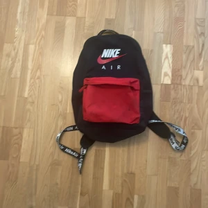 Svart och röd ryggsäck från Nike Air - Säljer en snygg svart ryggsäck från Nike Air med röd frontficka och vita logotyper på både väskan och axelremmarna. Perfekt för skola eller träning, med justerbara remmar och praktiska fack.