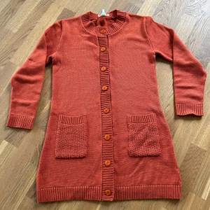 Rostfärgad stickad cardigan från Cissi och Selma - Lång, rostfärgad stickad cardigan med stora knappar framtill och två framfickor. Ribbstickade muddar vid ärmslut, hals och knappslå. Perfekt för lager-på-lager och enkel att styla.