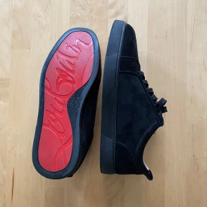 Svarta sneakers från Christian Louboutin - Säljer ett par svarta sneakers från Christian Louboutin i mocka med klassisk röd sula och dekorativa detaljer på ovandelen. Skorna har rund tå och platt sula. Kommer med originalkartong och dustbag. Pris är ej hugget i sten