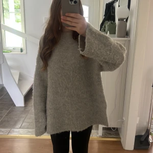 Oversized grå/beige stickad tröja - Supermjuk och oversized grå stickad tröja med lång ärm och slits i sidan. Tröjan har en kort polo vid halsen. Passformen är loose och den är riktigt skön att mysa i under kyliga dagar.
