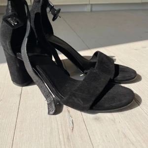 Svarta sandaler med klack från Vagabond - Stilrena svarta sandaler från Vagabond med bred rem över tårna och justerbart spänne runt ankeln. Skorna har en stadig blockklack och är tillverkade i mocka med detaljer i skinn. 