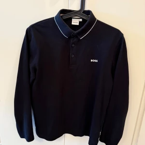 Svart långärmad pikétröja från BOSS - Snygg svart långärmad pikétröja från BOSS med klassisk krage och tre knappar framtill. Vit piping på kragen och diskret BOSS-logga broderad på bröstet. Perfekt för en stilren och clean look.