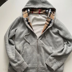 Grå hoodie från Burberry med dragkedja - Snygg grå hoodie från Burberry med klassiskt rutigt foder i huvan. Tröjan har dragkedja, snörning i huvan och två stora fickor framtill. Mjuk insida i fleece och ribbade muddar. Perfekt för dig som gillar stilrena och exklusiva plagg.