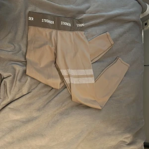 Beige leggings från Stronger -  Original pris ca 550kr! Nästan aldrig använda. Snygga beige leggings från Stronger med hög midja och bred resår med logga. De har två vita randiga detaljer med prickigt mönster på ena benet. Perfekta för träning eller chill. Materialet är stretchigt och känns mjukt mot huden. Pris kan diskuteras💗