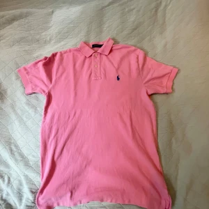 Rosa piké från Polo Ralph Lauren - Säljer en klassisk rosa pikétröja från Polo Ralph Lauren i storlek S. Tröjan har korta ärmar, krage och knäppning med två knappar framtill. Ikoniska blå loggan broderad på bröstet. Perfekt för en fräsch och sportig look. Om du vill starta en ny sak att ha rosa och inte vara som alla andra här är ett tips, denna piké är också en perfekt sommar present till någon som har barn i 13-15 års åldern 