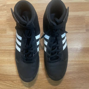 Svarta Adidas boxningsskor med vita ränder - Säljer ett par svarta Adidas boxningsskor med klassiska vita ränder på sidorna. Skorna har snörning, hög modell med kardborreband upptill och gummisula med bra grepp. Perfekta för träning eller match.