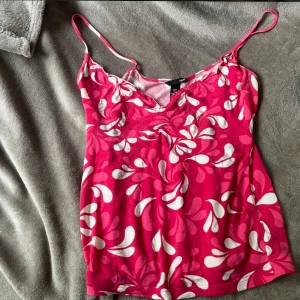 Rosa och vit blommig linnetopp H&M - Snygg rosa linnetopp från H&M med vita blommiga mönster och tunna axelband. Toppen har en lätt v-ringning och är figurnära med stretchigt material, perfekt för varma dagar. Färgen är starkt rosa med vita detaljer som ger en fräsch och somrig känsla.