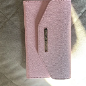 Rosa plånbok från iDeal of Sweden - Snygg ljusrosa plånbok från iDeal of Sweden med stilren design och magnetlås. Plånboken har plats för kort och mobil, och är gjord i ett tåligt syntetmaterial med en elegant metallogo på framsidan.