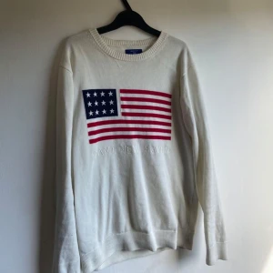 Vit stickad tröja från Gant - Vit stickad tröja från Gant med broderad amerikansk flagga på bröstet och texten 'GANT NEW HAVEN' under. Tröjan har rund halsringning och långa ärmar, perfekt för en klassisk och avslappnad stil.