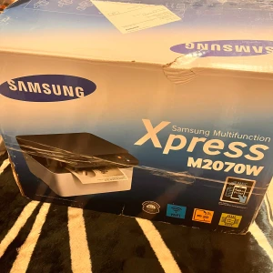 Skrivare - Bilderna visar en Samsung Xpress M2070W multifunktionsskrivare i originalkartong. Produkten är en skrivare, inte en bok, och passar perfekt för studenter eller unga vuxna som behöver skriva ut, kopiera och skanna hemma. Enkel anslutning via WiFi och mobilapp.