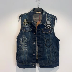 Jeansväst  - Cool mörkblå jeansväst med slitna detaljer, dödskalle-patch på bröstet och texten 'NO MORE WAR' på axeln. Västen har knappar framtill, två bröstfickor och två sidofickor. Perfekt för en edgy streetstil.
