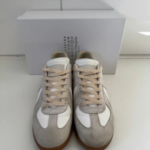 Maison Margiela gats - Snygga sneakers från Maison Margiela med vit och ljusgrå ovandel i skinn och mocka, beige snörning och brun gummisula. Klassisk och stilren design som passar till många outfits. Kvitto finns !