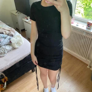 Svart ribbad kortklänning med snörning - Säljer en svart kortklänning med ribbad struktur och snörningsdetaljer på sidorna. Klänningen har tight passform, korta ärmar och rund halsringning. Perfekt för dig som gillar en enkel men snygg stil.