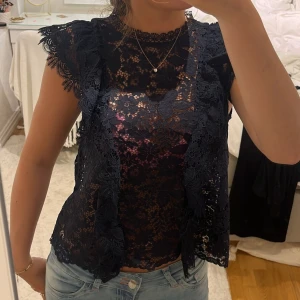 Mörkblå spetsblus - Säljer en Mörkblå blus i spets med vackra blommönster och volangdetaljer vid axlarna. Blusen har rund halsringning och är kortärmad. Använd en gång. 